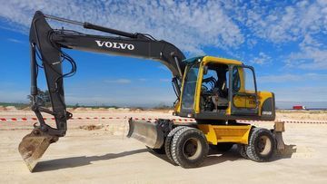 Excavadora Volvo amarilla con brazo negro sobre terreno arenoso bajo un cielo azul.