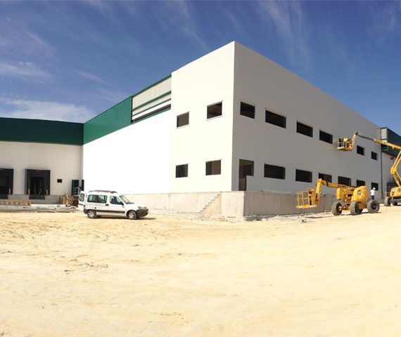 Gran edificio industrial blanco y verde en construcción; vehículos sobre suelo arenoso.