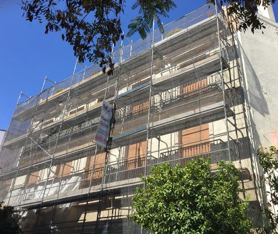 Fachada de edificio cubierta con andamios y redes durante la renovación. Cielo azul y vegetación.