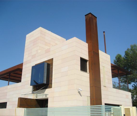 Edificio moderno de dos pisos con fachada de piedra arenisca, gran ventanal y alta chimenea de metal contra un cielo azul.
