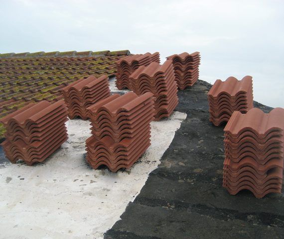 Pilas de tejas rojas sobre un tejado plano cubierto de negro con un tejado parcialmente de tejas en el fondo.
