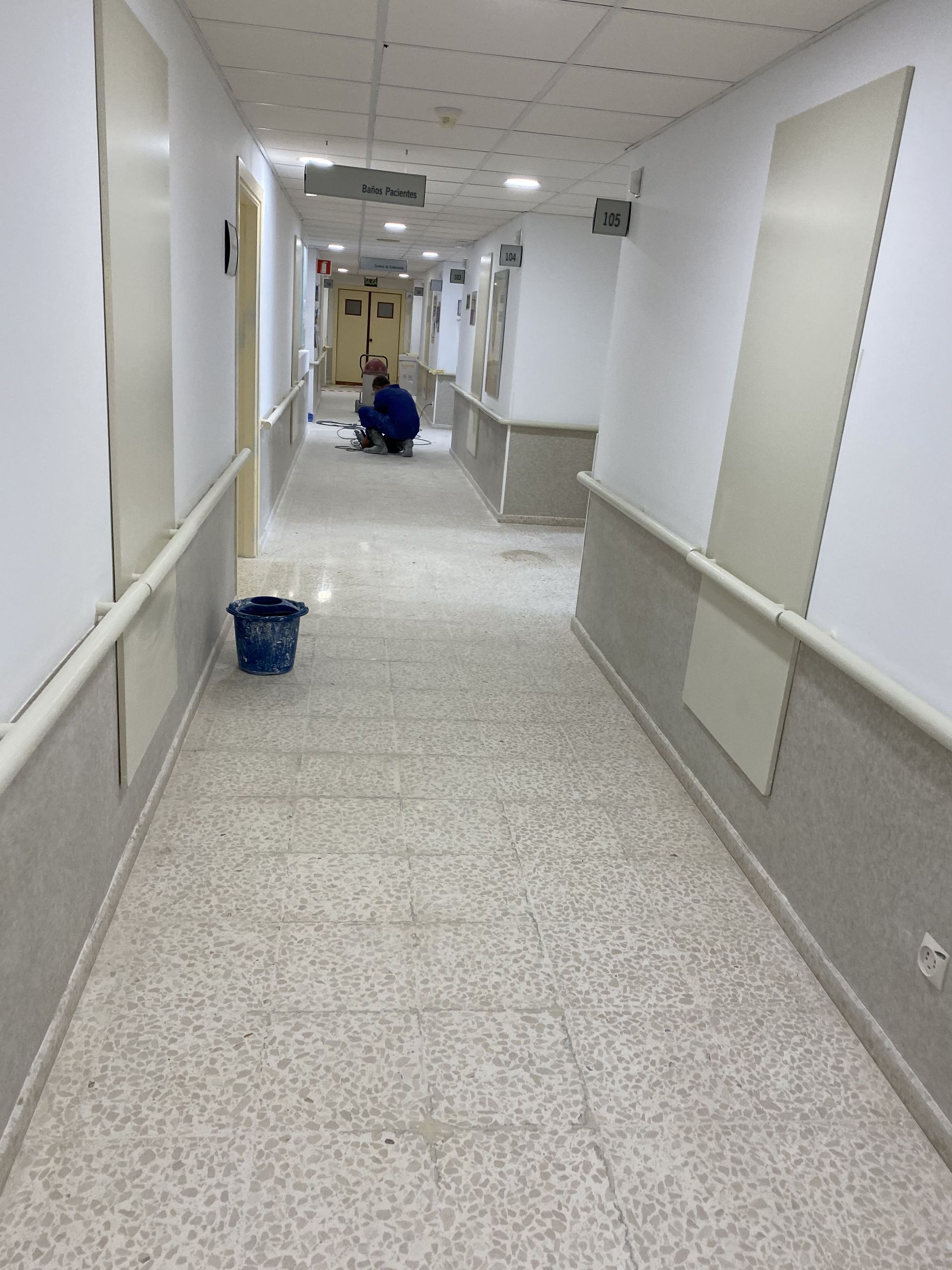 Pasillo largo con piso de baldosas de color claro, paredes y molduras blancas y una persona trabajando en la distancia.