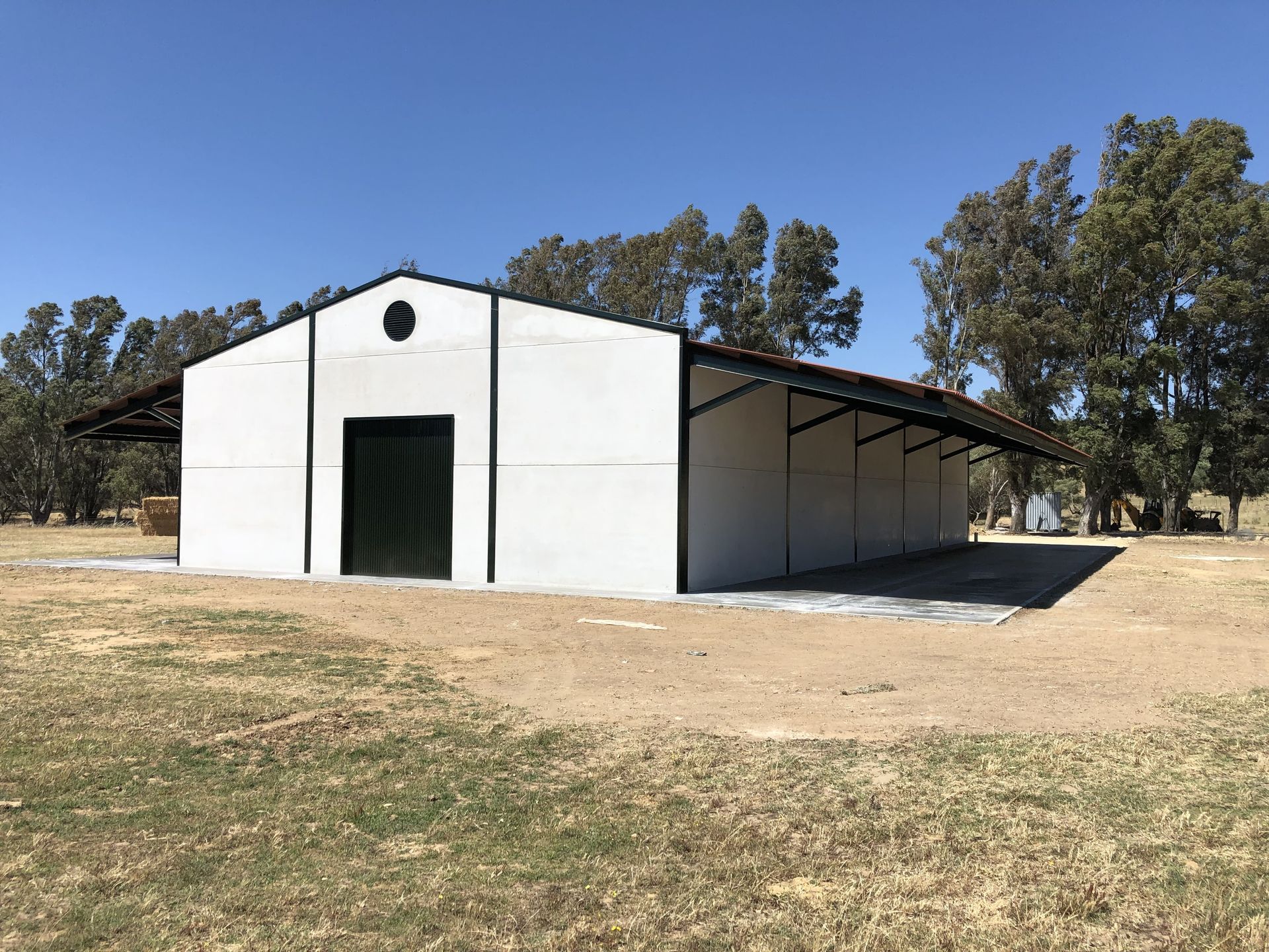 Granero blanco con detalles negros, puerta verde y alero cubierto en un campo de hierba bajo un cielo azul.