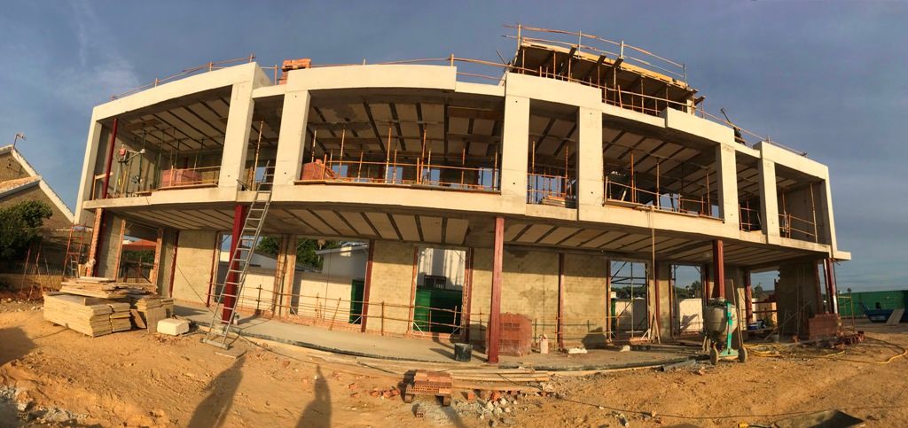 Edificio en construcción con paredes de hormigón y espacios abiertos. Ubicado en un terreno de tierra, parcialmente soleado.