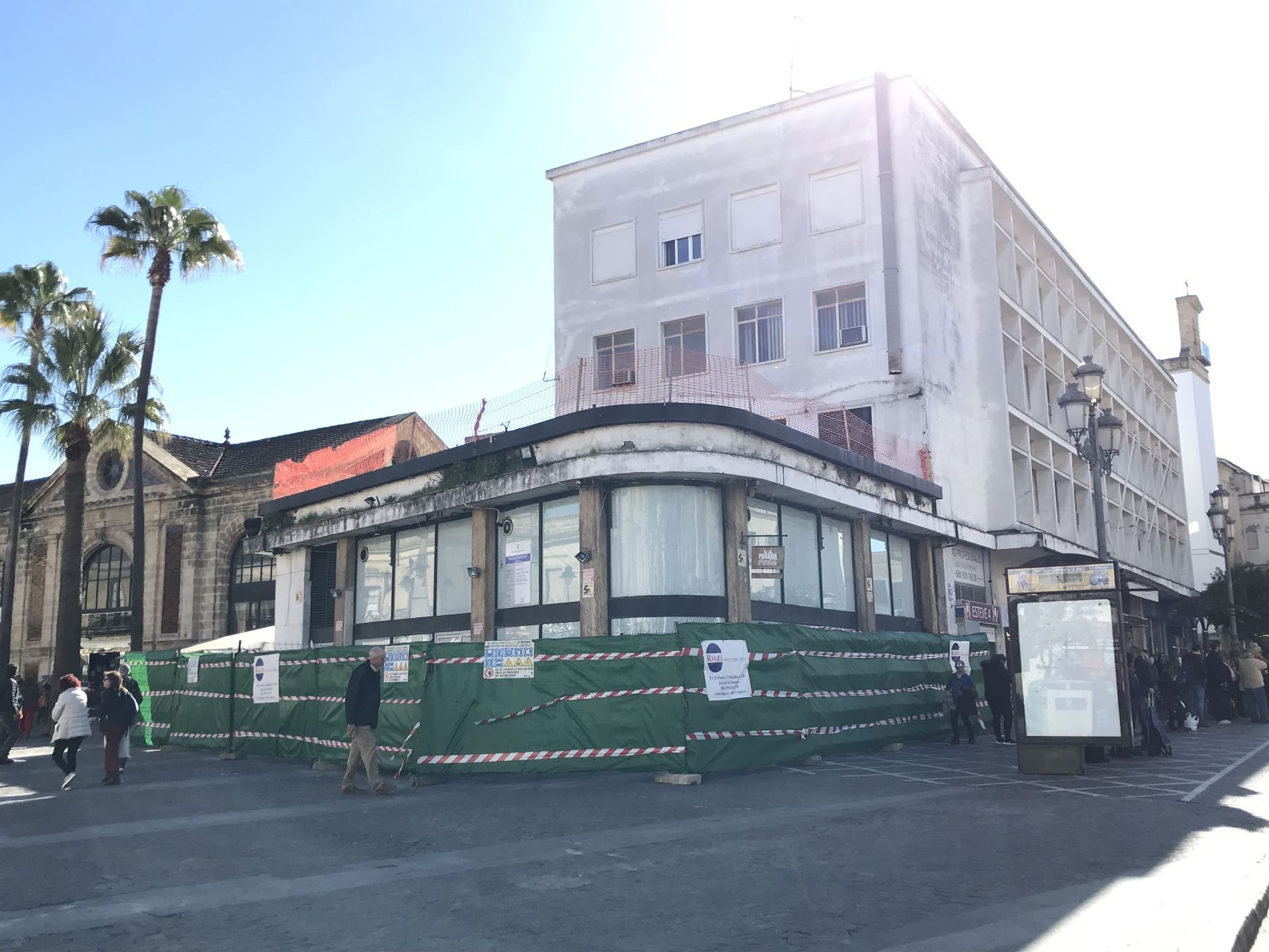 Edificio en construcción, con barreras verdes, junto a un edificio alto y una iglesia en un día soleado.