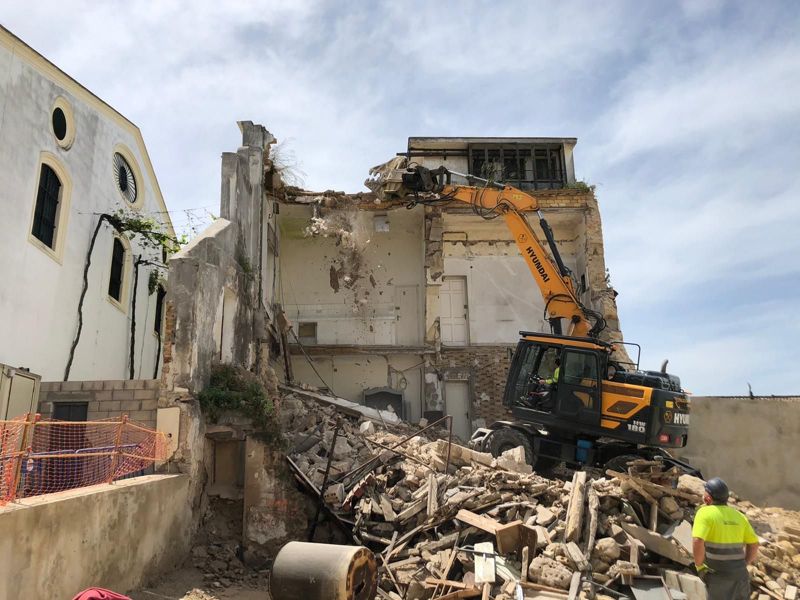 Un obrero con chaleco amarillo observa cómo una excavadora demuele un edificio. Escombros y escombros rodean la zona.