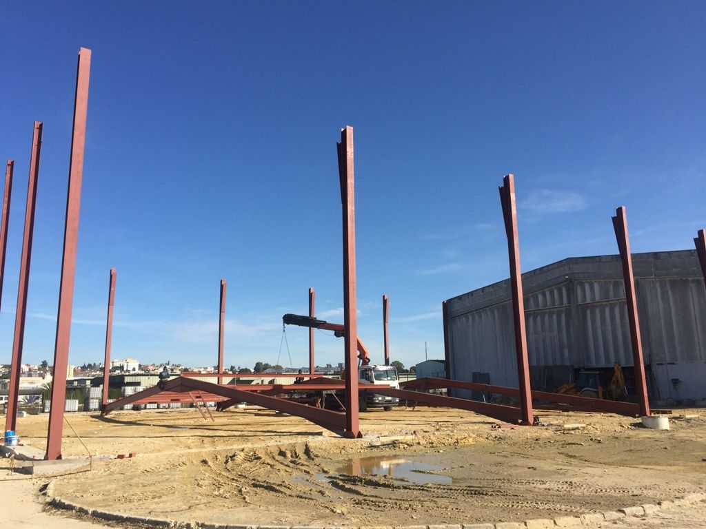 Obra en construcción con vigas de acero contra un cielo azul claro; al fondo hay un edificio de hormigón.