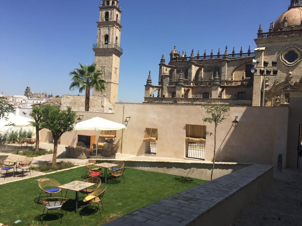 Patio exterior con mesas y sillas sobre césped, iglesia y torre al fondo bajo un cielo azul.