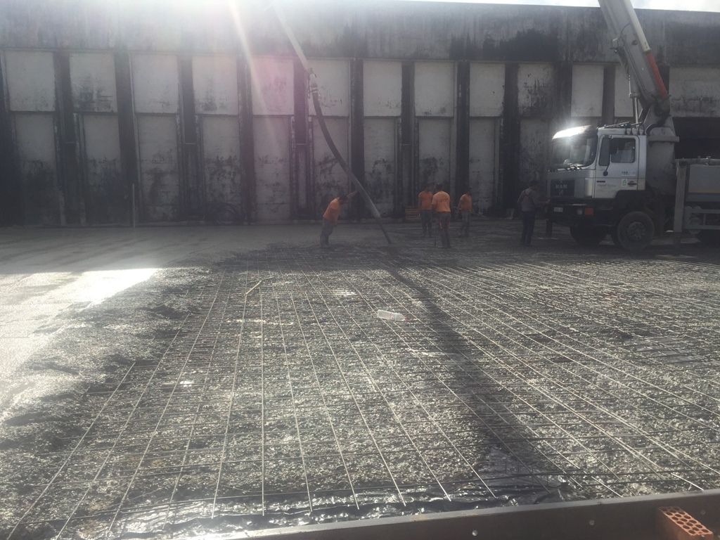 Obreros de la construcción vierten hormigón sobre una rejilla de refuerzo, con un camión hormigonera y un edificio al fondo.