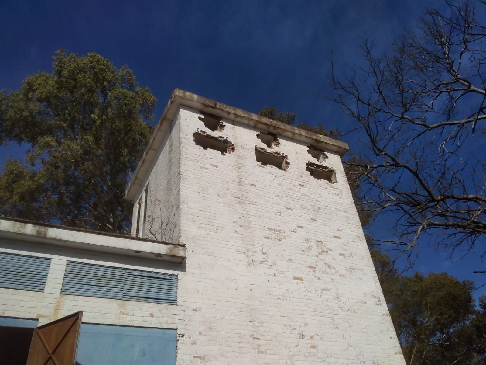 Torre de ladrillo blanco con aberturas de ventilación bajo un cielo azul claro, árboles al fondo.