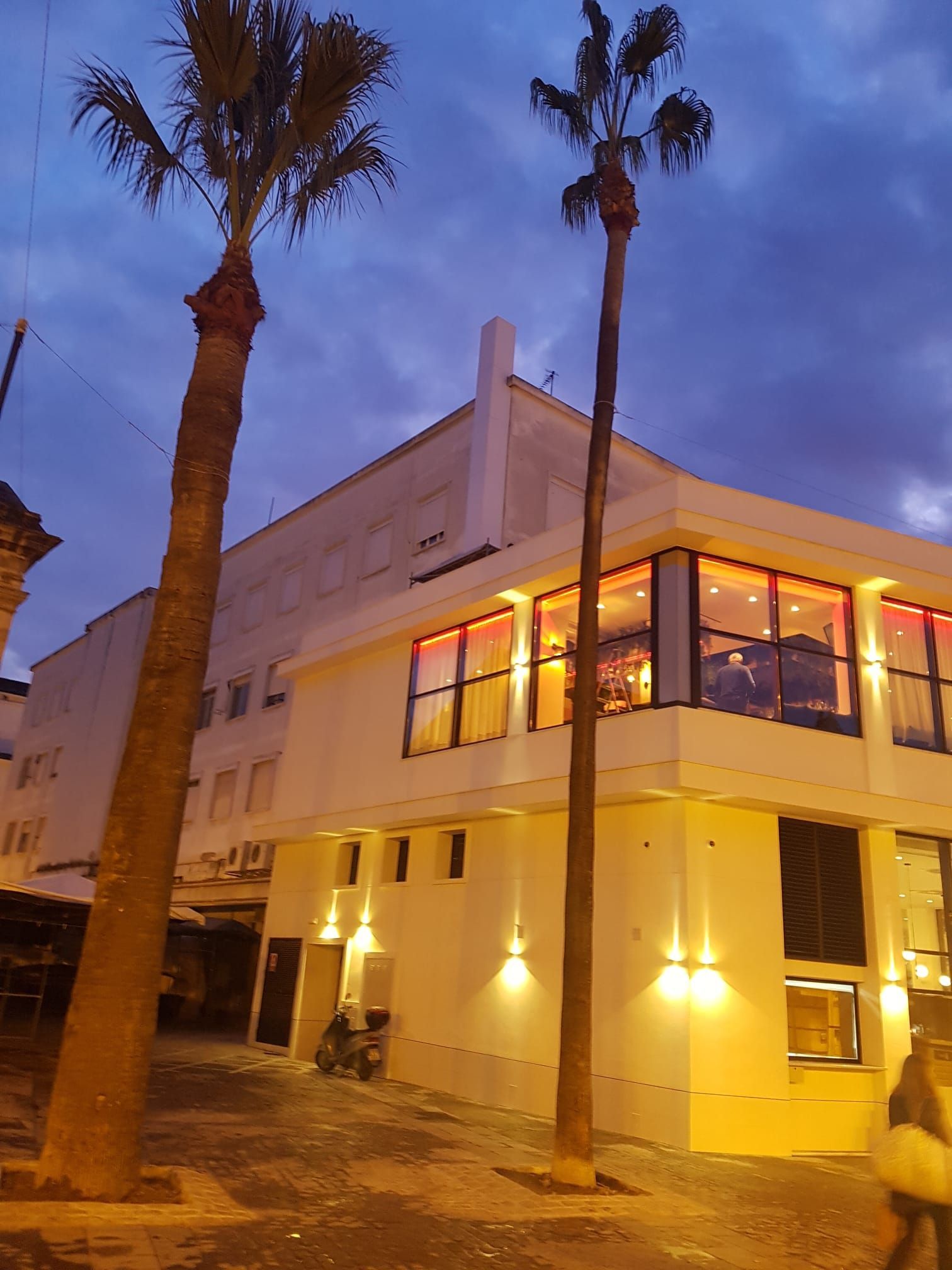 Un edificio blanco al atardecer con palmeras, ventanas iluminadas y luces exteriores.