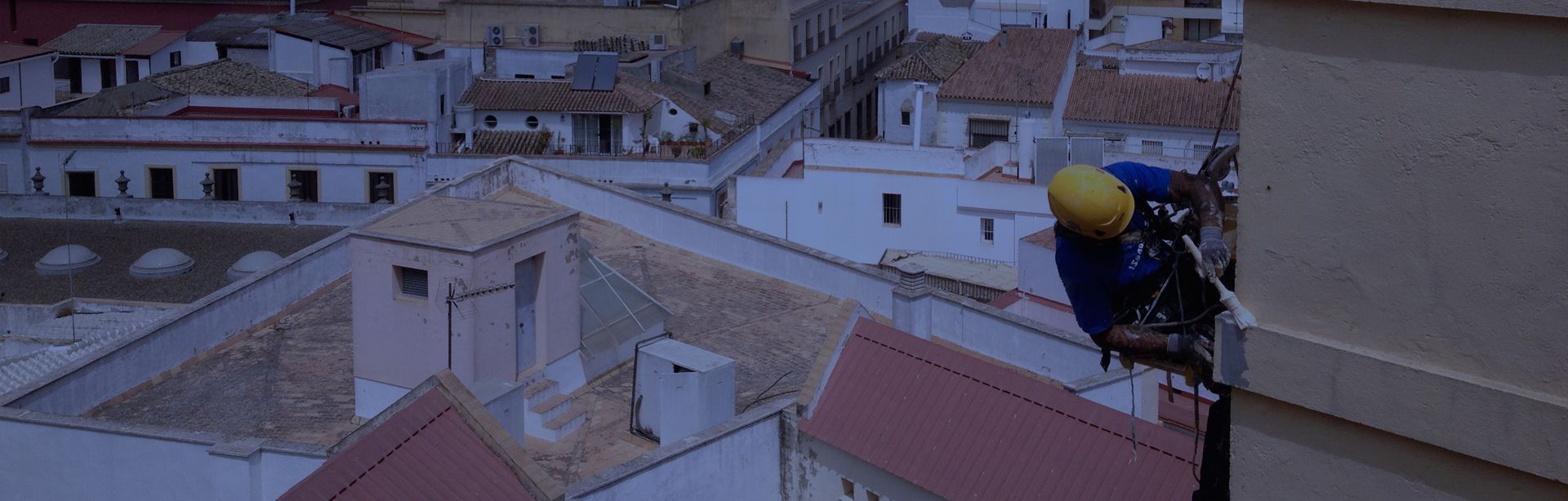 Una persona con un mono azul y un casco amarillo sube a un edificio, con un paisaje urbano de fondo.