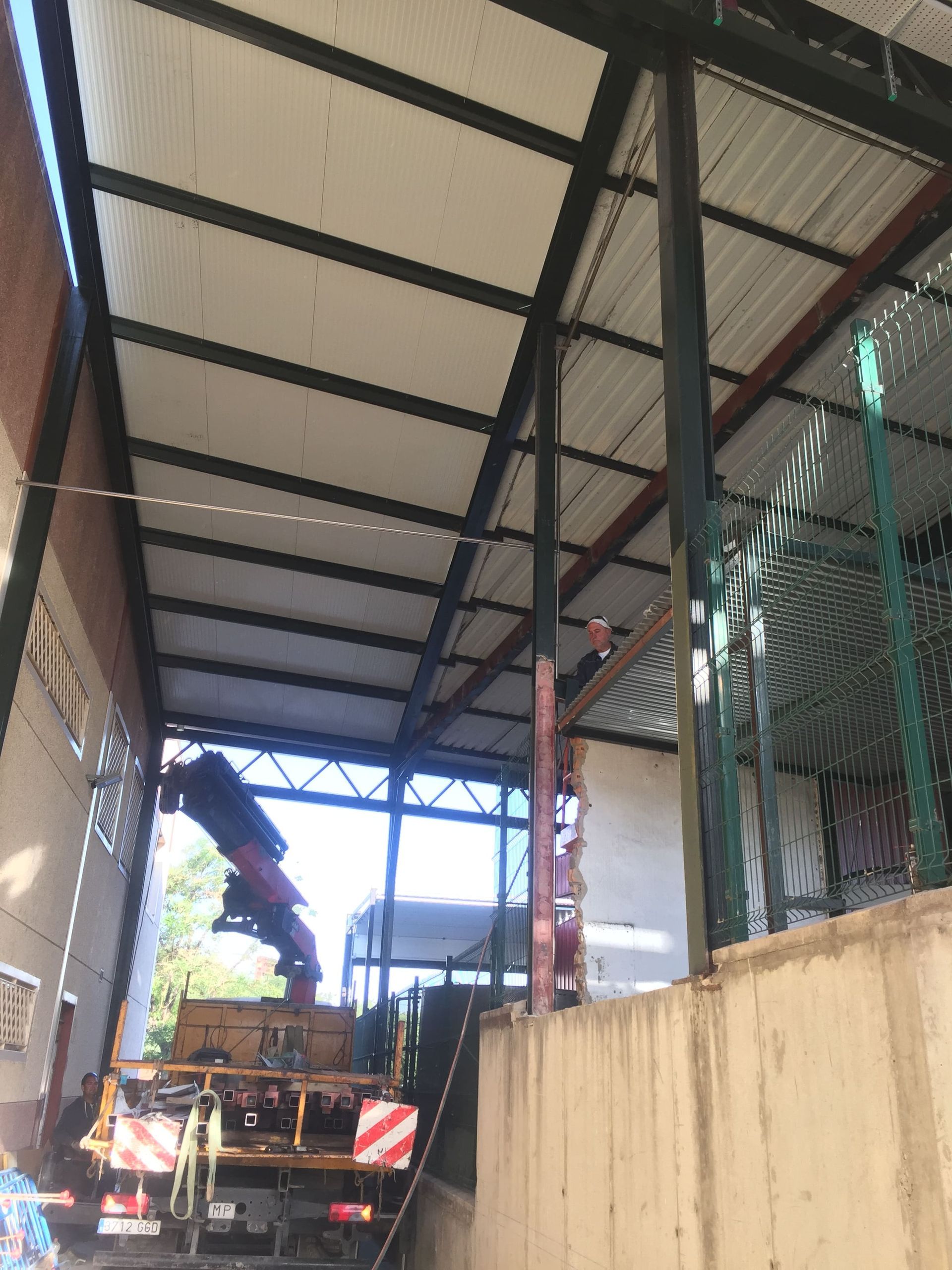 Una grúa eleva un equipo bajo una estructura metálica. Muro de hormigón, edificio a la izquierda y valla a la derecha.