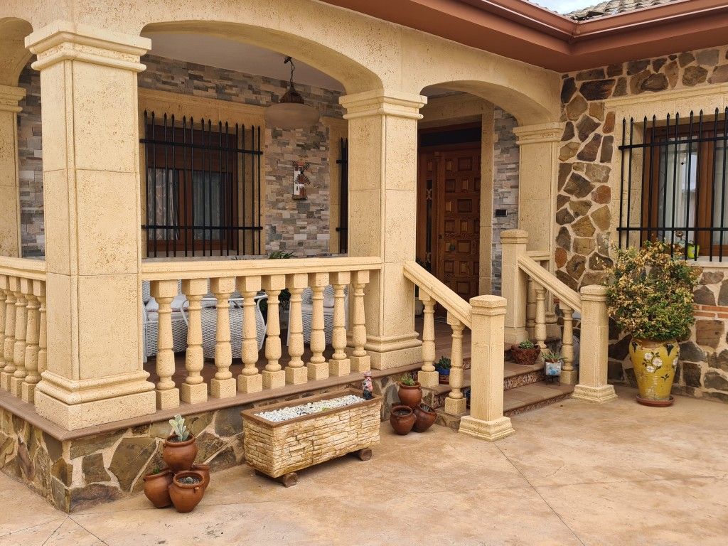 Porche de piedra con columnas, entrada arqueada, puerta de madera y plantas en macetas.