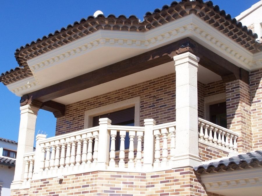Fachada de una casa de ladrillo con barandillas blancas en el balcón y tejas de terracota bajo un cielo azul claro.