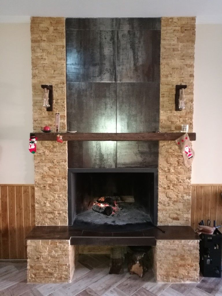 Chimenea de piedra con revestimiento de azulejos oscuros, repisa de madera y fuego encendido en su interior.