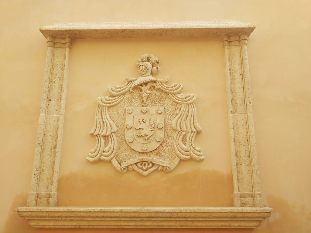 Placa con escudo de armas tallada en piedra sobre una pared beige, enmarcada por columnas y molduras decorativas.