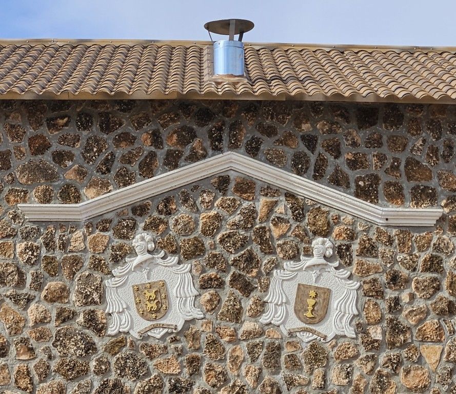 Muro de piedra con placas decorativas con escudos de armas bajo un tejado de tejas y una chimenea metálica de ventilación.