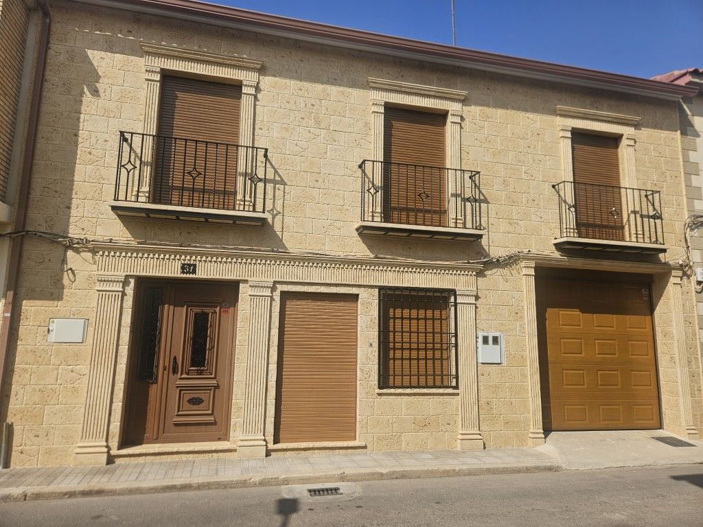Fachada de piedra con tres balcones, puertas de madera y puerta de garaje en una calle soleada.
