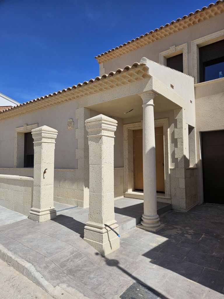 Entrada de piedra a la casa con tres columnas y un porche cubierto bajo un cielo azul brillante.