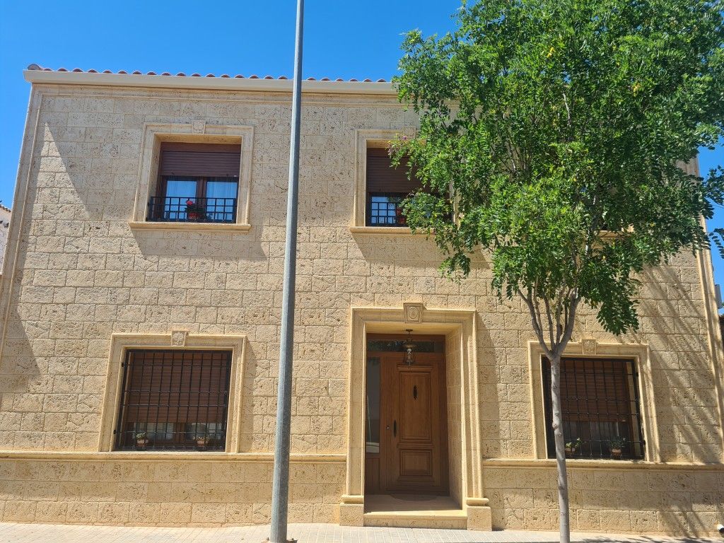 Edificio de piedra de dos plantas con ventanas enrejadas, puerta central de madera y un árbol delante bajo un cielo azul.