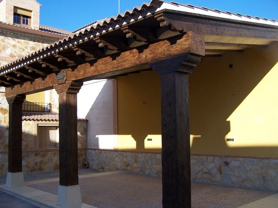 Patio cubierto de piedra con vigas de madera oscura, pared amarilla y techo de tejas bañado por la luz del sol.