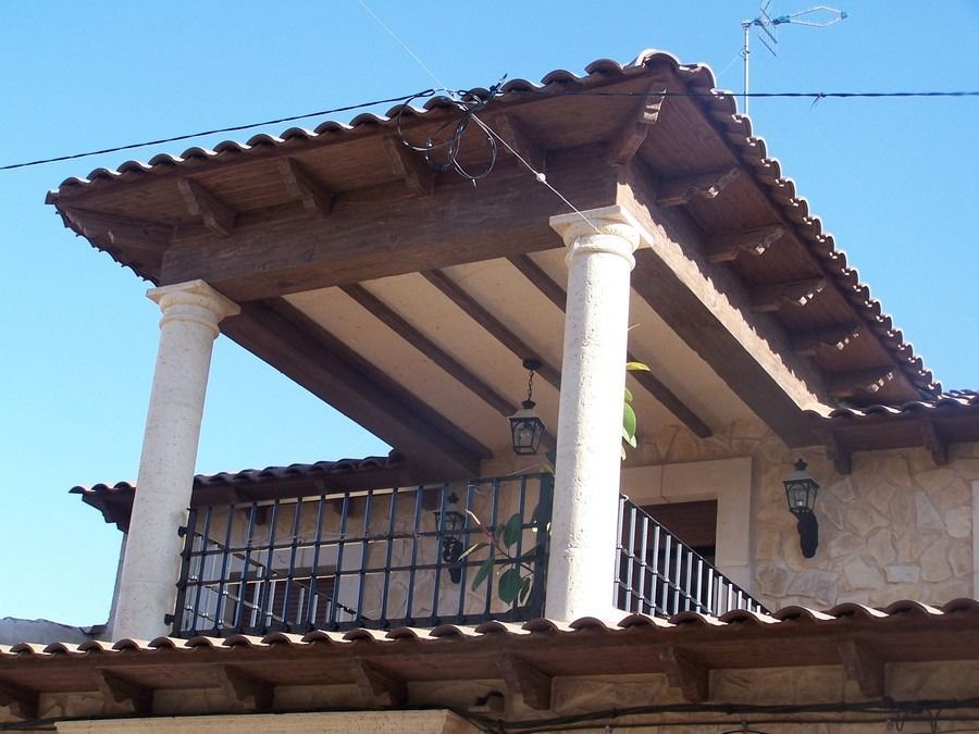 Balcón de piedra cubierto con columnas blancas y techo de tejas de terracota bajo un cielo azul.