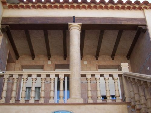 Balcón beige con columnas de piedra, vigas de madera y techo de tejas contra una pared de estuco.