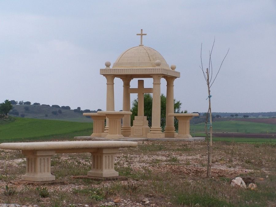Pequeño santuario de piedra con cúpula y cruz en un campo rural, con un banco delante y cielo nublado.