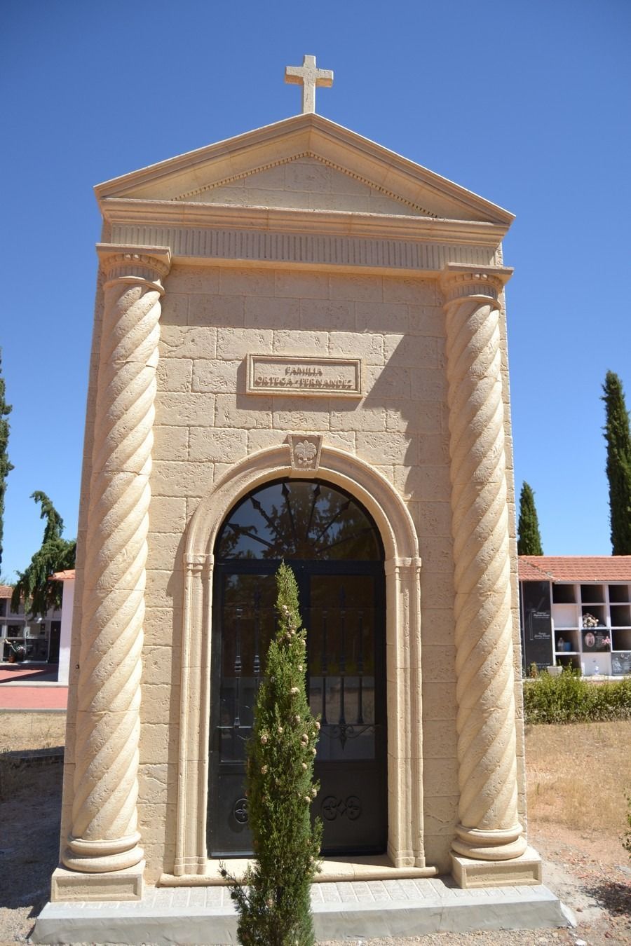 Pequeña entrada de una capilla de piedra con una cruz en la parte superior y un ciprés delante, bajo un cielo azul.