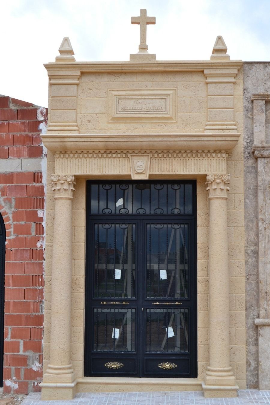 Elegante portal de piedra beige con puertas dobles negras y una cruz en la parte superior.