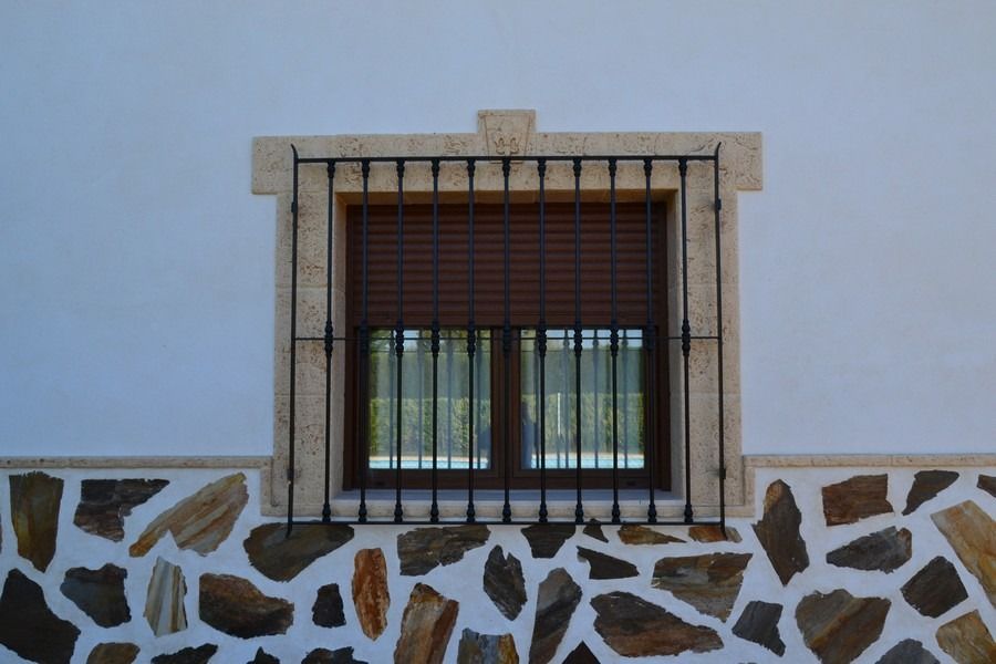 Ventana enrejada con marco de piedra beige sobre una pared blanca decorada con motivos de piedra marrón.