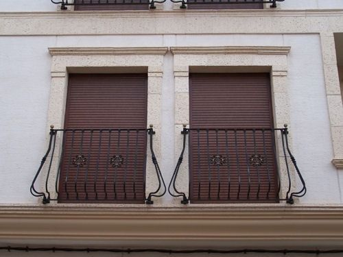 Dos contraventanas marrones cerradas en la fachada de un edificio beige con barandillas negras.