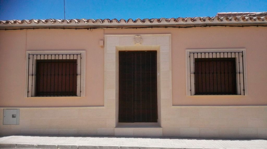 Vista frontal de la fachada de una casa de color beige con una puerta central y dos ventanas enrejadas bajo un cielo azul.