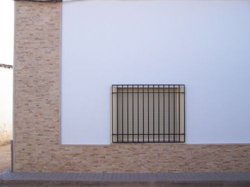 Pared blanca con una pequeña ventana enrejada sobre una base de ladrillo, junto a un sendero pavimentado.