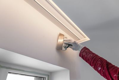 Eine Hand mit grauem Handschuh streicht mit einem kleinen Pinsel eine weisse Deckenecke in der Nähe eines Fensters.