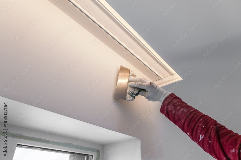 Eine Hand mit grauem Handschuh streicht mit einem kleinen Pinsel eine weisse Deckenecke in der Nähe eines Fensters.