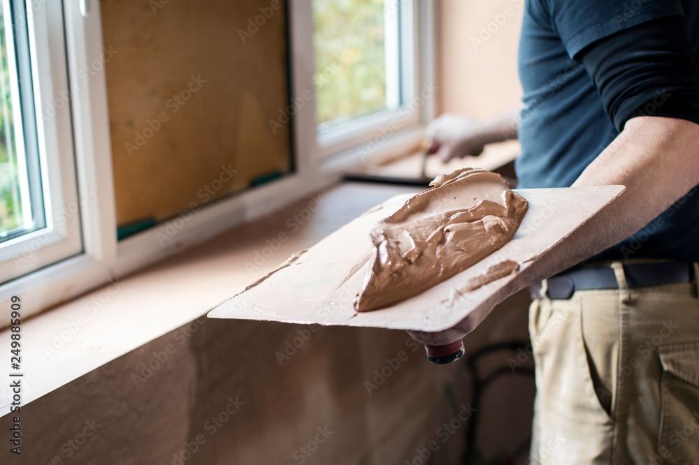 Eine Person hält eine mit Schlamm bedeckte Kelle neben einem Fenster und verputzt eine Innenwand.