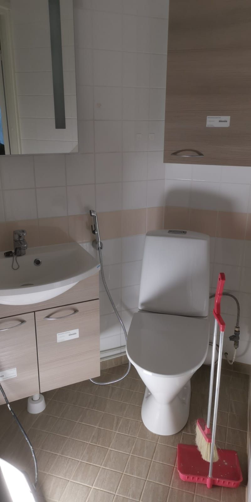 Kylpyhuone, jossa on valkoinen wc-pönttö, pesuallas sekä punainen rikkalapio ja luuta. Seinät ovat laatoitettu ja lattia on ruskea.