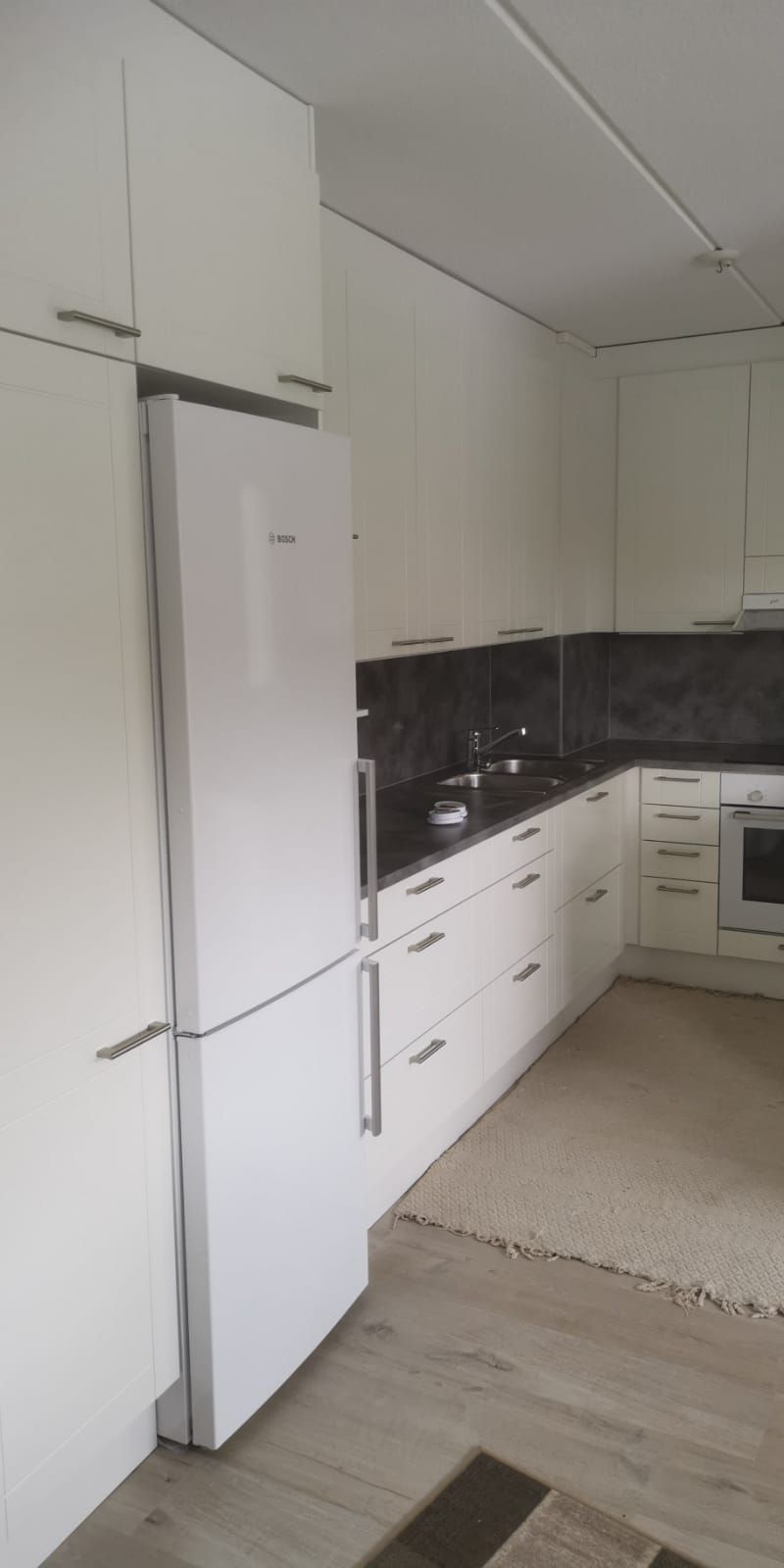 Valkoinen keittiö, jossa jääkaappi ja kaapit; puulattia.