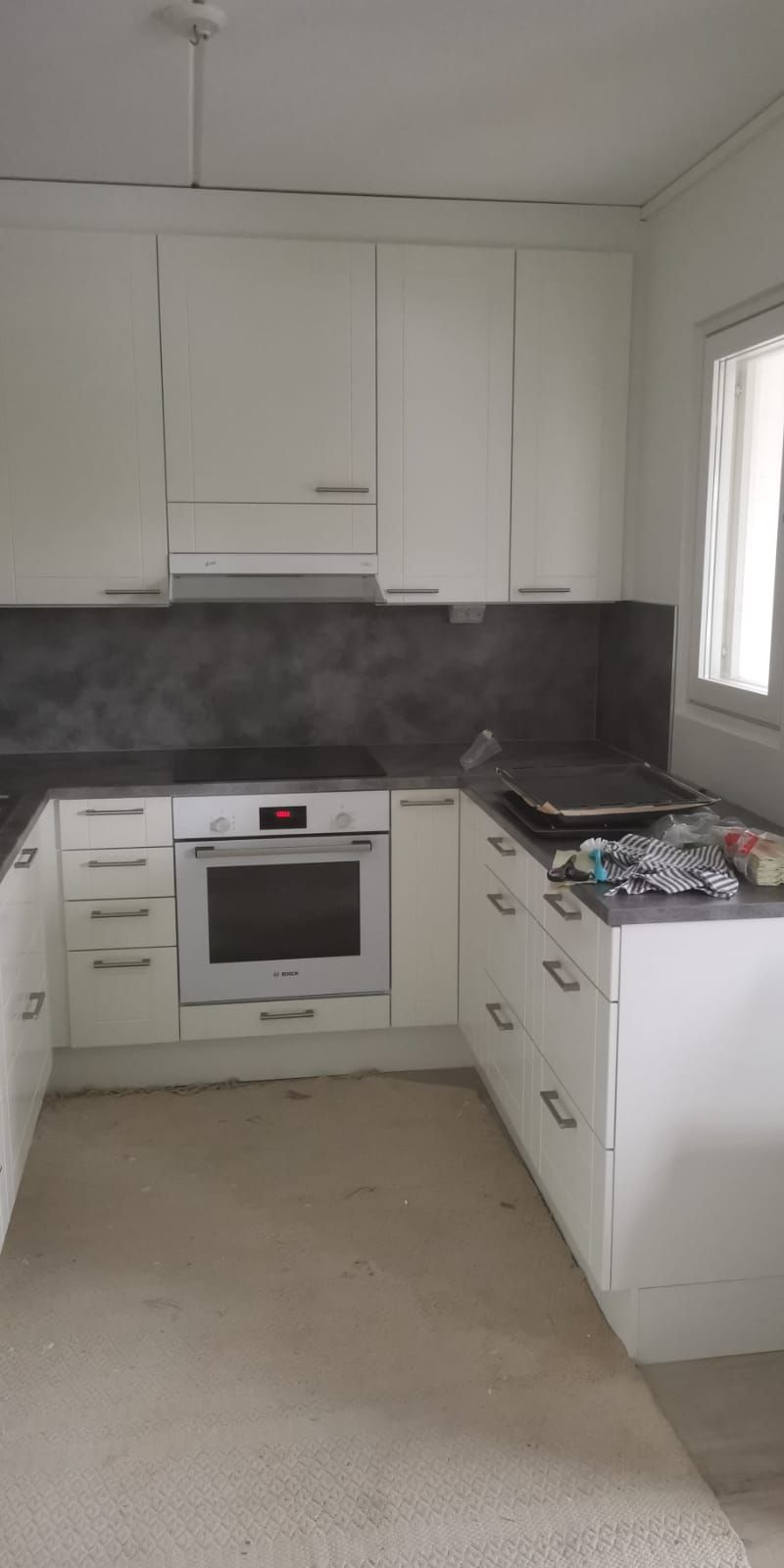 U-muotoinen valkoinen keittiö, jossa harmaa välitilalevy ja ruostumattomasta teräksestä valmistettu uuni. Luonnonvaloa ikkunasta.