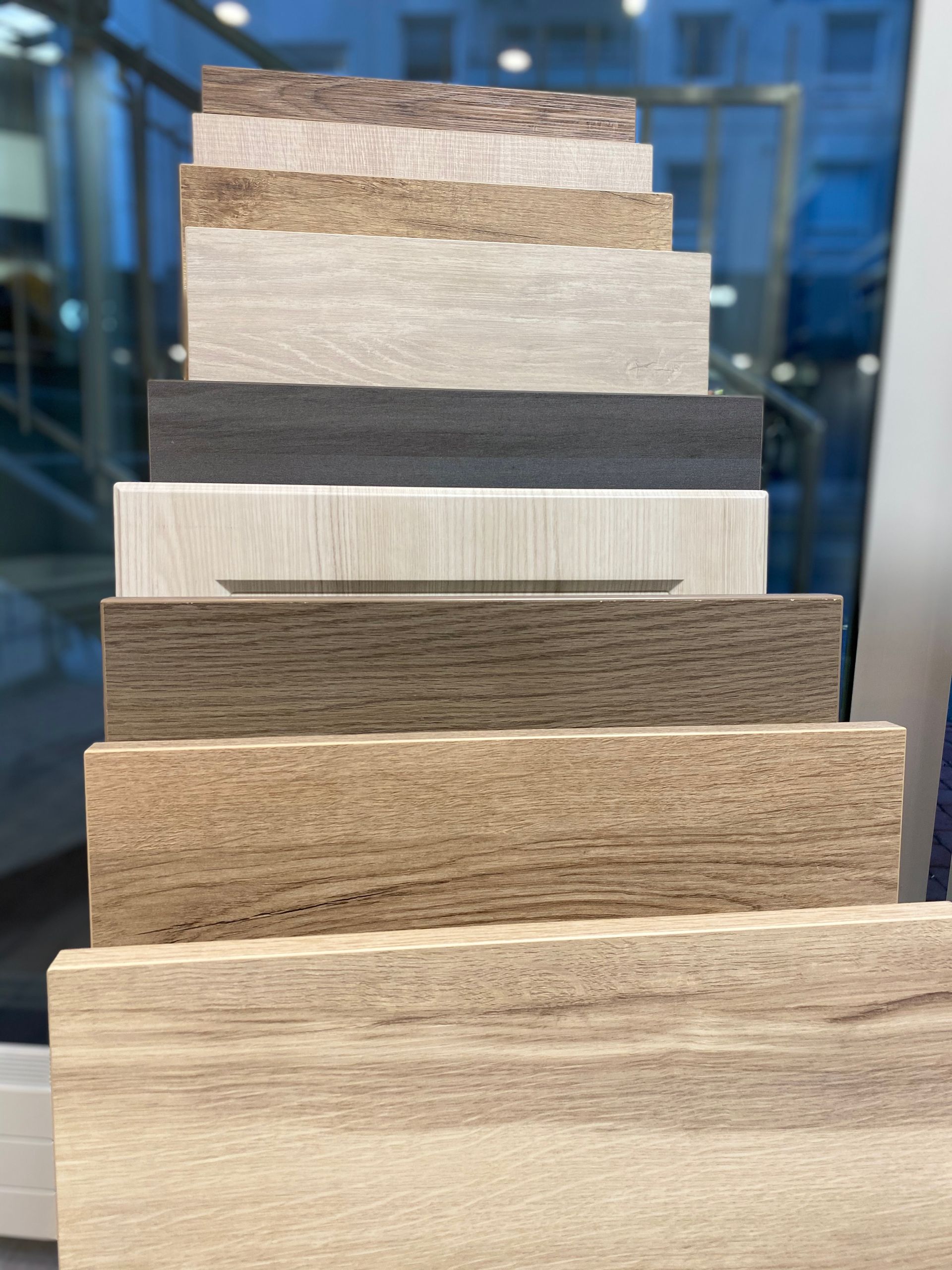 Pinottu kokoelma puukuvioisia ja kivijäljitelmiä sisältäviä lattianäytteitä ikkunaa vasten; erilaisia värejä ja tekstuureja.