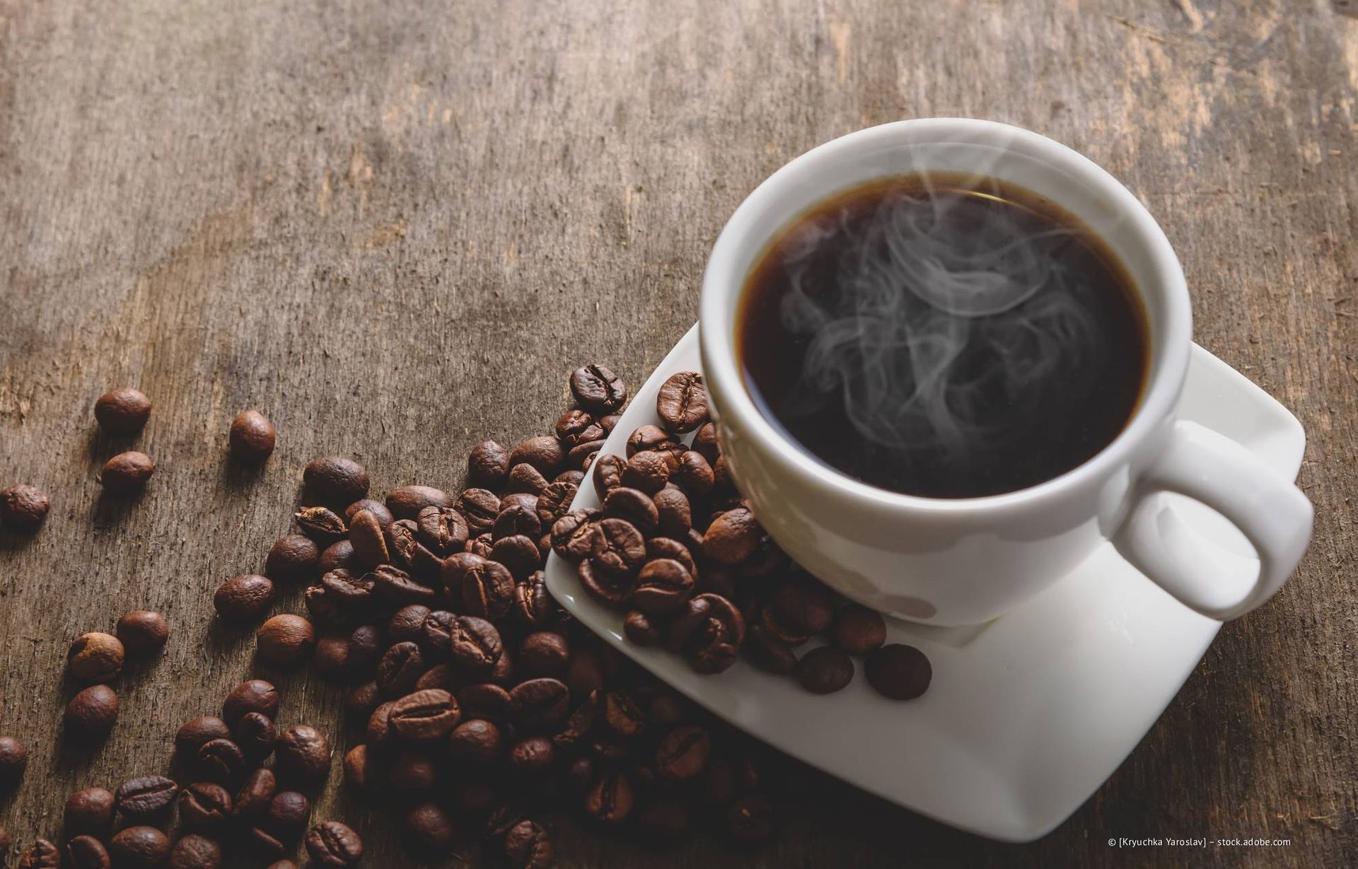 Dampfender Kaffee in einer weißen Tasse auf einer Untertasse, umgeben von gerösteten Kaffeebohnen auf einer Holzoberfläche.