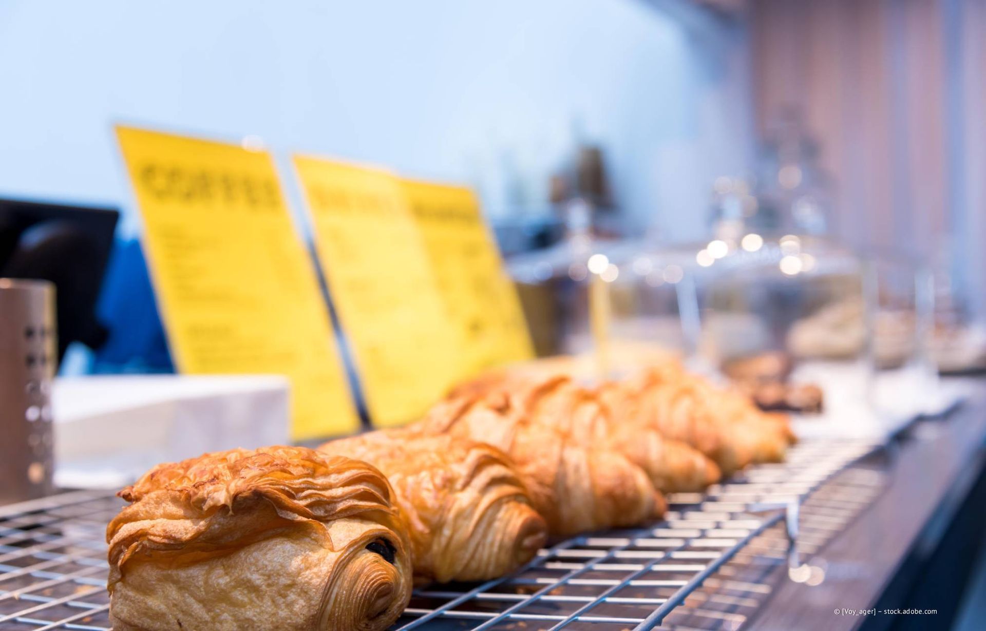 Croissants und Gebäck auf einem Metallregal in einer Bäckerei, im Hintergrund eine gelbe Kaffeekarte.