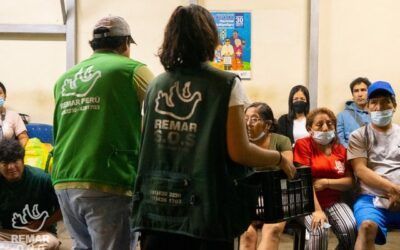 Dos personas con chalecos verdes se dirigen a un público sentado en una sala que parece un aula.