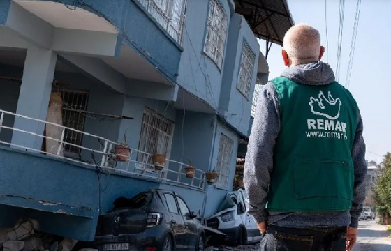 Una persona con un chaleco verde inspecciona un edificio azul derrumbado con coches aplastados debajo.