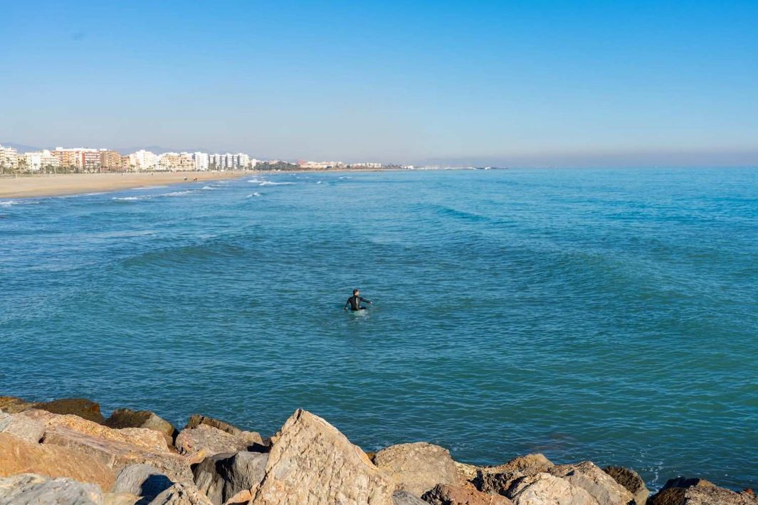 Costa rocosa con vistas a un mar azul tranquilo, con un surfista solitario y edificios costeros a lo lejos.