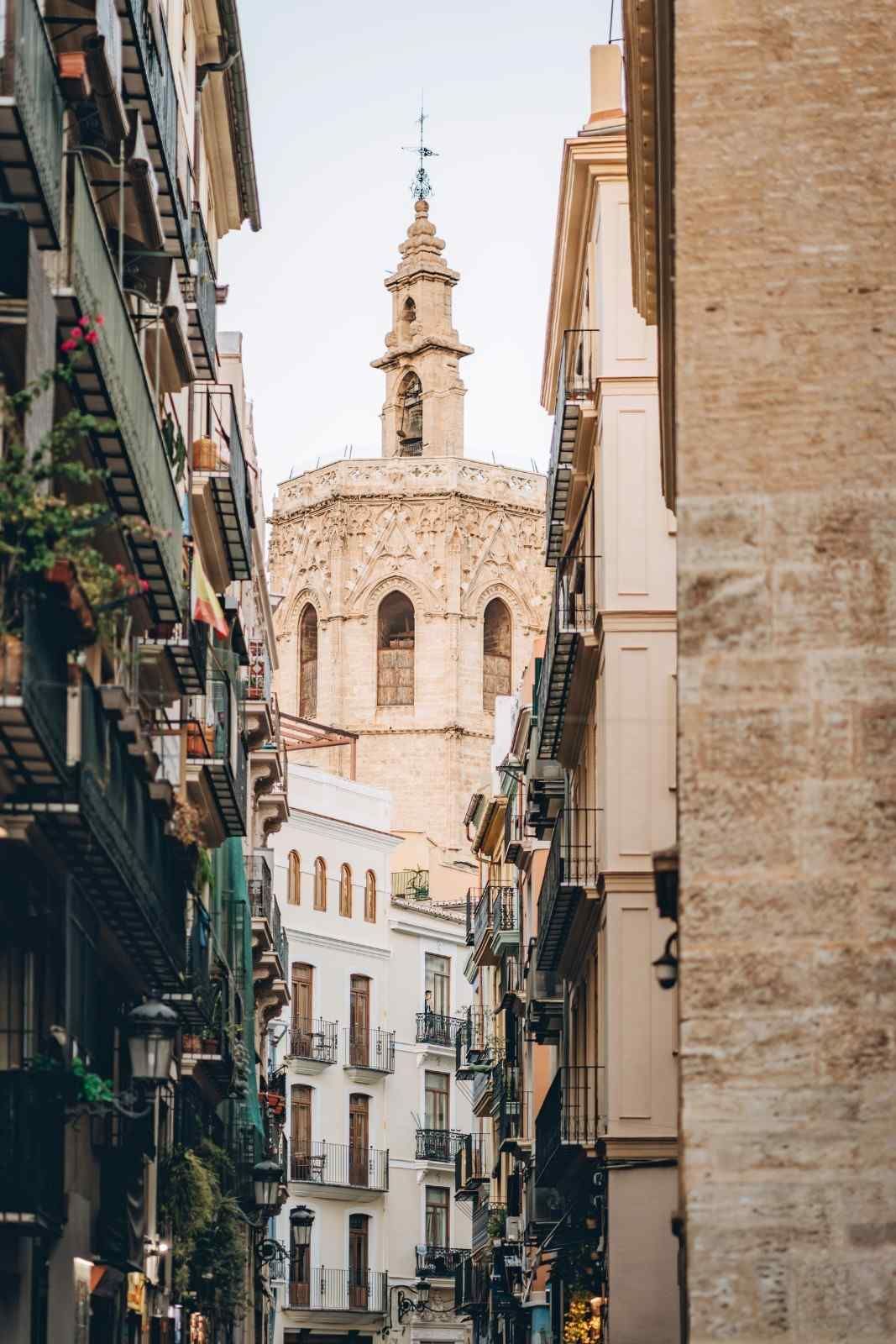 Calle estrecha de estilo europeo con balcones, que conduce a una clara torre de iglesia de piedra bajo un cielo despejado.