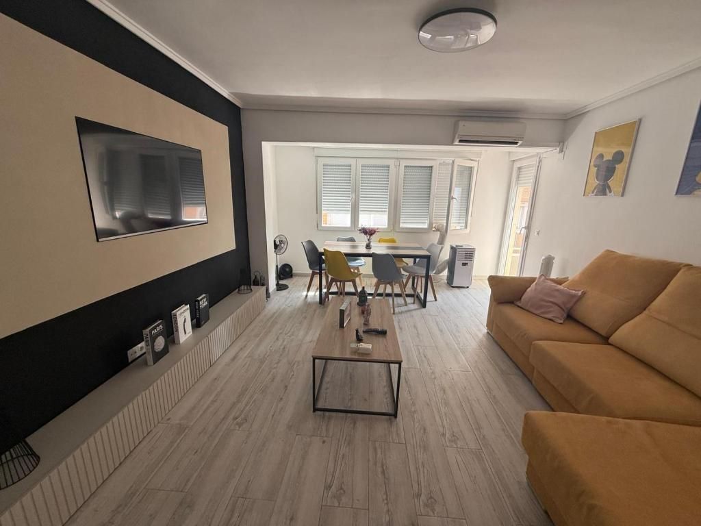 Sala de estar moderna con sofá color canela, televisor montado en la pared, mesa de centro y zona de comedor al fondo.
