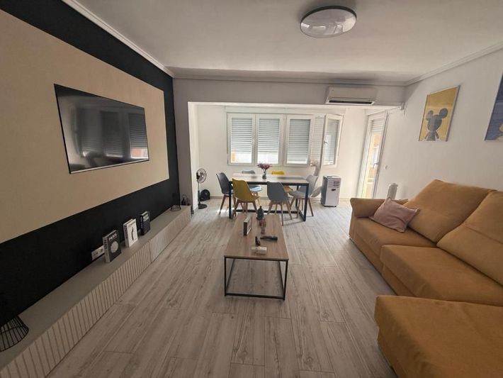 Sala de estar moderna con sofá color canela, televisor montado en la pared, mesa de centro y zona de comedor al fondo.